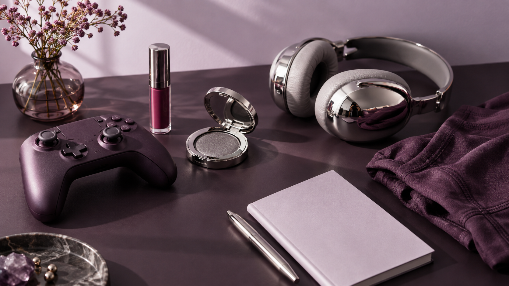 A plum game controller, berry lip tint, pewter compact and chrome headphones on a creative desk.